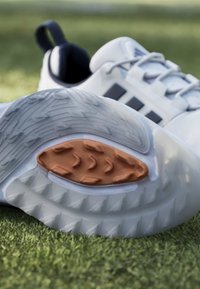Chaussures de sport blanches avec semelles en caoutchouc texturées, une chaussure montrant un coussinet adhérent orange, posées sur une surface d'herbe verte en extérieur.