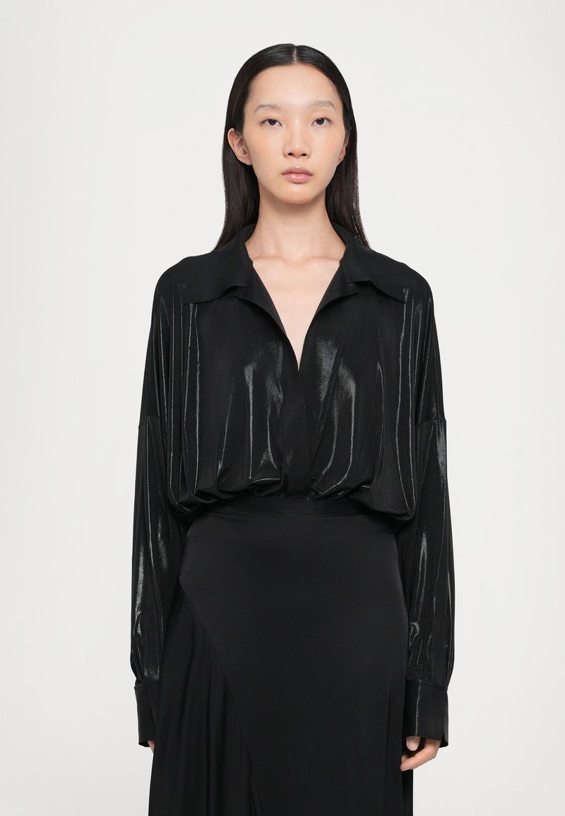 Black, long-sleeve blouse with a V-neck, glossy texture, loose fit, and wide collar. Worn over a black skirt, creating a sleek silhouette.