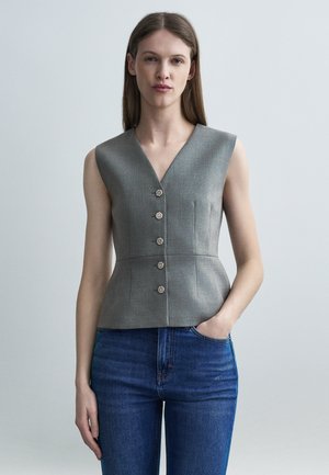 Nő, aki ujjatlan szürke, testhezálló mellényt visel öt díszgombbal és kék farmert, egy sima világos háttér előtt áll.