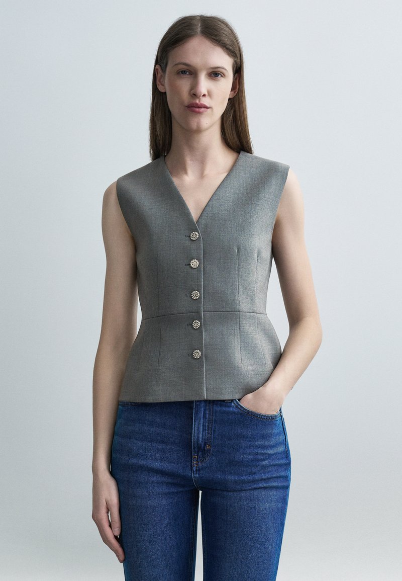 Γυναίκα που φοράει γκρι αμάνικο εφαρμοστό γιλέκο με πέντε διακοσμητικά κουμπιά και μπλε τζιν παντελόνι, στέκεται μπροστά σε ένα απλό φωτεινό φόντο.