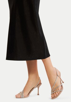 Nogi w cielistych sandałach na wysokim obcasie z paskami ozdobionymi kryształkami, jedna stopa lekko uniesiona, w czarnej spódnicy midi na jednolitym tle.