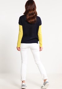 Marinblå och gul tvåfärgad tröja med korta ärmar, kombinerad med vita jeans och grå sneakers, sedd bakifrån.