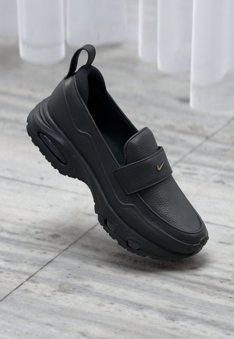 Chaussure en cuir noire sans lacets avec une semelle épaisse et un petit logo Nike, présentée sur un sol en marbre gris clair avec des rideaux blancs en arrière-plan.