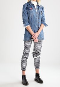 Osoba v modré džínové bundě s nášivkami, v černo-bíle kostkovaných kalhotách, černých kotníkových botách a držící malou stříbrnou kabelku.