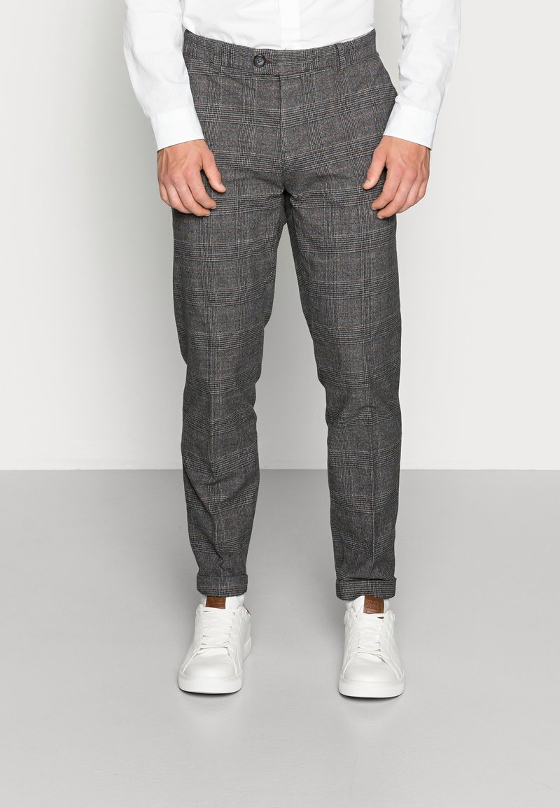 Hombre vestido con pantalones de cuadros grises y zapatillas blancas de pie frente a un fondo claro y liso.