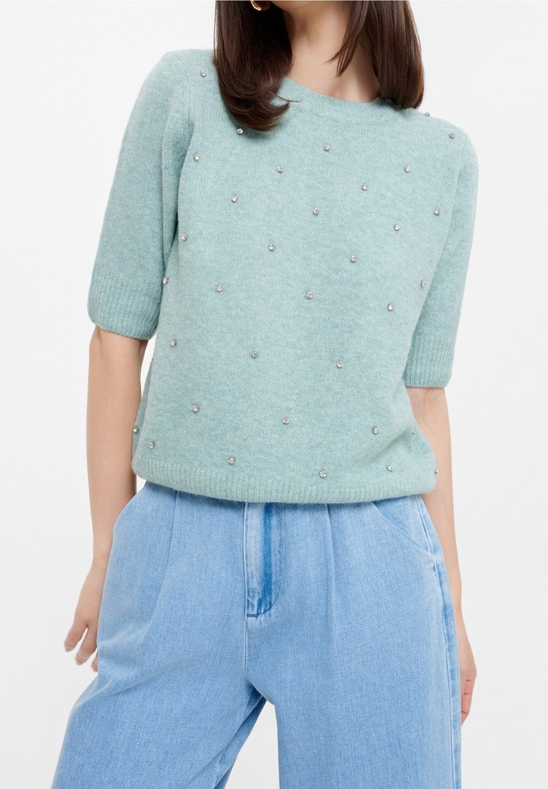 Femme portant un pull tricoté bleu clair à manches courtes avec de petites perles en décoration, associé à un pantalon en denim clair taille haute.