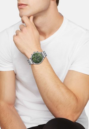 Silver stainless steel watch with a dark green face, three subdials, and simple hour markers, worn on a bare wrist.