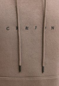 Καφέ φούτερ με κουκούλα από μαλακό ύφασμα με υφή στην επιφάνεια. Διαθέτει τύπωμα λογότυπου, πλεγμένα κορδόνια και μεταλλικά διακοσμητικά στοιχεία.