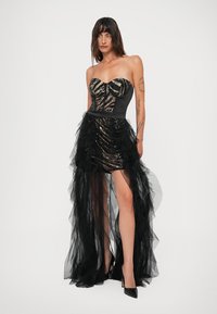Model u strapless crnoj i zlatnoj mini haljini od šljokica s prozirnim crnim tilom, u crnim visokim potpeticama, stoji naslonjena na bijelu pozadinu.