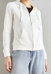 Felpa bianca con zip e cordoncini, polsini e orlo in maglia, dotata di tasche frontali e una texture morbida, indossata sopra una maglietta scura con jeans blu.