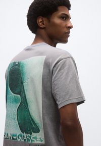 Γκρι κοντομάνικο μπλουζάκι με μεγάλο πράσινο σχέδιο γυαλιών ηλίου στην πλάτη και μπλε ραφές στο λαιμό.