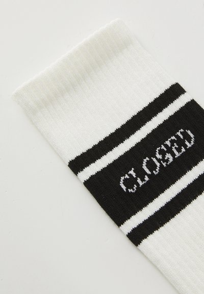 Chaussette blanche avec un haut côtelé, présentant des accents rayés noir et blanc et le mot "CLOSED" dans une police en gras sur un fond noir.