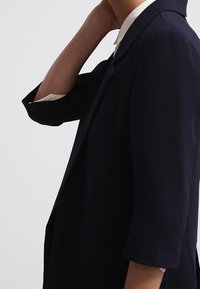 Námořnický modrý blazer s přiléhavým střihem, tříčtvrtečními rukávy, klopovým límcem a jednou náprsní kapsou. Hladká textilie.