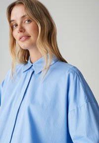Camicia oversize azzurro chiaro con colletto classico, chiusura con bottoni e tessuto liscio, caratterizzata da spalle scese e una vestibilità rilassata.