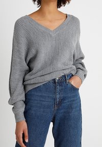 Femme portant un pull gris ample en maille à col en V, rentré dans un jean bleu taille haute avec une main dans la poche.