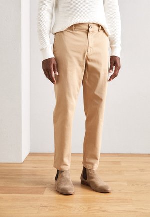 Mężczyzna w beżowych spodniach, beżowych zamszowych butach Chelsea i białym, teksturowanym swetrze stoi na drewnianej podłodze na tle jednolitej ściany.