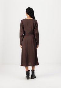 Robe marron à manches longues avec une silhouette fluide, taille cintrée et texture lisse. Associée à des bottines noires.
