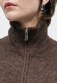 Καφέ, ριγωτό πουλόβερ με φερμουάρ και ψηλό γιακά. Μαλακή υφή και εφαρμοστή γραμμή, με έμφαση στη λαιμόκοψη και τις λεπτομέρειες στο πάνω μέρος του σώματος.