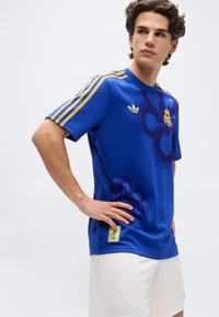 Young man in blue Adidas sports shirt with yellow stripes and white shorts, standing with hands on hips against plain background.