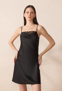 Robe nuisette en satin noir avec un ourlet en dentelle au niveau du col, dotée de fines bretelles et d'une silhouette décontractée qui tombe juste au-dessus du genou.