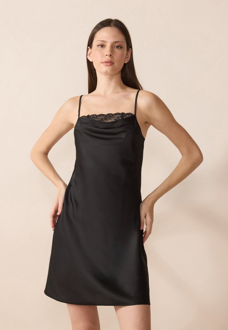 Robe nuisette en satin noir avec un ourlet en dentelle au niveau du col, dotée de fines bretelles et d'une silhouette décontractée qui tombe juste au-dessus du genou.