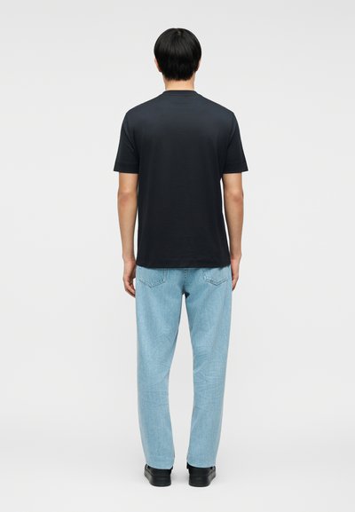 Άνδρας που φοράει μαύρο κοντομάνικο μπλουζάκι, ανοιχτό μπλε τζιν και μαύρα παπούτσια, στέκεται με την πλάτη γυρισμένη σε απλό φόντο.