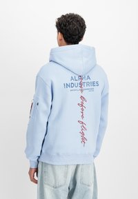 Világoskék kapucnis pulóver, amelyen az "ALPHA INDUSTRIES" logó és piros kiegészítő szöveg található a hátsó részen. Puha anyagból készült, bordázott mandzsettákkal.