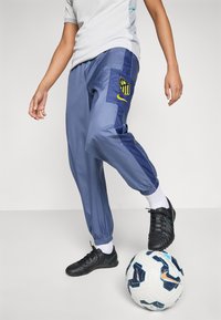 Une personne portant un pantalon de sport bleu et des crampons noirs équilibre un ballon de soccer Nike blanc et bleu avec son pied.