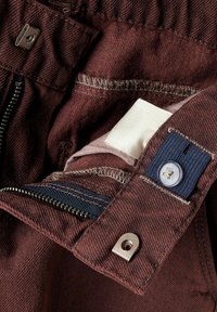 Pantaloni bordeaux con interno a zip, cuciture a contrasto e etichetta in tessuto. Presentano un pulsante blu e dettagli in metallo.