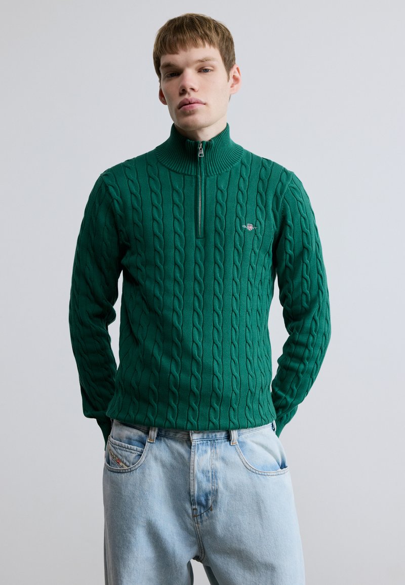 Green knitted sweater with a high collar and zip closure, featuring a cable pattern and small embroidered logo. Worn with light blue jeans.