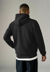 Zwarte textuur hoodie met een capuchon, voorzien van een geribde onderrand en manchetten. Draagt over een wit shirt, gezien vanaf de achterkant.