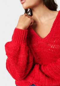 Czerwony sweter wykonany na drutach z głębokim dekoltem w kształcie V, z teksturowanym, otwartym splotem i ściągaczami na mankietach. Miękki materiał i luźny krój.
