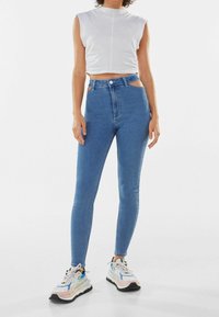 Vaqueros de mezclilla azul de talle alto con detalles recortados en las caderas, combinados con un top corto blanco ajustado y zapatillas multicolores.