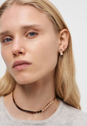 Blonde Frau mit blauen Augen, die quadratische goldene Ohrringe und eine passende goldene Halskette mit quadratischen Gliedern trägt, hellgraues Oberteil, neutraler Hintergrund.