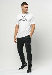 Mann iført hvit T-skjorte med svart logo, svarte joggebukser med logo, svarte joggesko, står med den ene hånden i lommen og ser til siden.