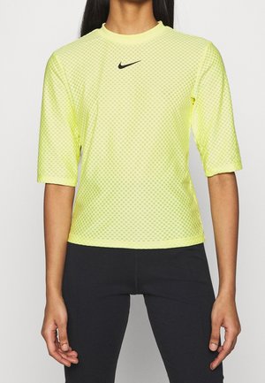 Kobieta ubrana w żółtą kratkę sportową koszulkę z krótkim rękawem z czarnym logo Nike i czarne legginsy, stojąca na jednolitym tle.