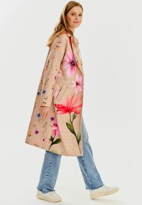 Kvetinový béžový kabát s ružovými a fialovými kvetinovými vzormi, dlhé rukávy a uvoľnený strih, skombinovaný so svetlomodrými džínsami a bielymi teniskami.
