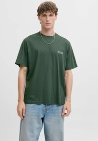 Tume roheline puuvillane t-särk, millel on lõdva istuvusega, lühikesed varrukad ja väike valge teksti logo vasakul rinnaosal. Kombineeritud helesiniste teksadega.