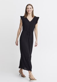 Robe maxi noire avec des manches à volants, décolleté en V, taille à cordon de serrage et ourlet évasé. Portée avec des sandales à talons vertes. Texture de tissu lisse.