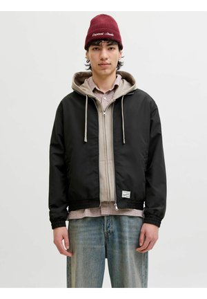 Junger Mann, der eine burgunderrote Mütze, einen beigen Hoodie, eine schwarze Zip-Up-Jacke, ein karierte Hemd und ausgewaschene blaue Jeans trägt, steht vor einem schlichten Hintergrund.