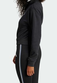 Felpa nera con colletto alto, polsini a coste e tasche laterali. Abbinata a leggings neri con una striscia verticale bianca.