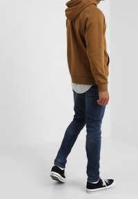 Man i brun hooded sweatshirt, blå jeans och svarta sneakers som går i ett vitt, tomt rum med enkla väggar och golv.