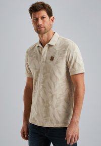 Polo beige clair avec un motif de feuilles discret, manches courtes, col classique et un petit écusson logo sur la poitrine.