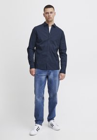 Camicia a maniche lunghe blu navy con bottoni, realizzata in cotone, abbinata a jeans di denim azzurro e sneakers bianche con strisce nere.