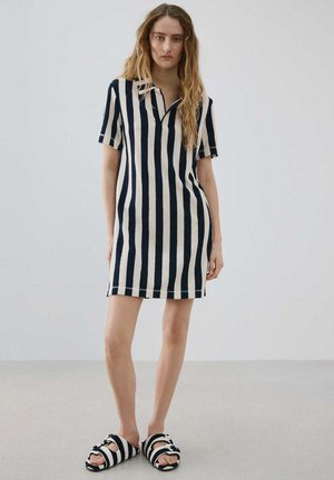 Joven mujer con vestido de manga corta a rayas verticales negras y blancas y sandalias a juego a rayas, de pie sobre un fondo liso.