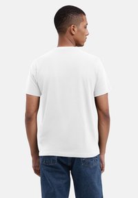 Weißes Baumwoll-T-Shirt mit kurzen Ärmeln, klassischem Rundhalsausschnitt und lockerer Passform, getragen von einer Person, die von hinten schaut, kombiniert mit dunkelblauen Jeans.