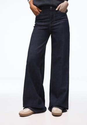 Personne portant un jean taille haute bleu foncé à jambes larges avec les mains dans les poches et des baskets beige clair sur fond blanc.