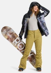 Snowboarder in giacca nera con un motivo di alberi colorati, maglietta beige e pantaloni gialli, tiene una tavola da snowboard grafica con il logo "ROXY".
