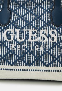 Niebieska i biała torba tkana z teksturowym wzorem, z logo "GUESS" wyhaftowanym białą nicią oraz skórzanymi uchwytami. Strukturalny design z akcentami.