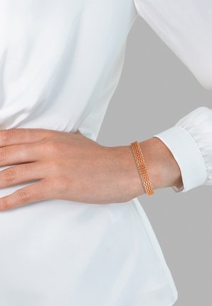 Mano di donna sul fianco che indossa una camicetta bianca a maniche lunghe e un bracciale in oro rosa lavorato al polso, su sfondo grigio.